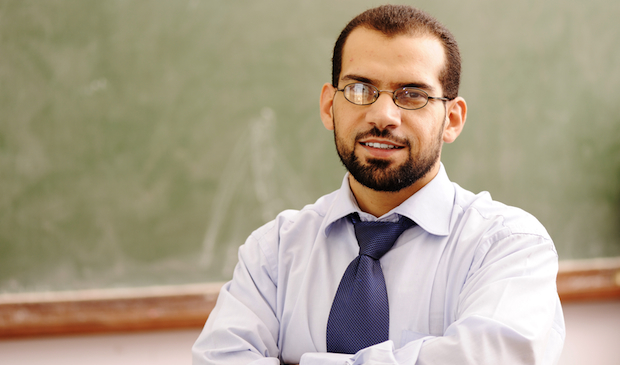 A confident male teacher stands in front of a chalkboard